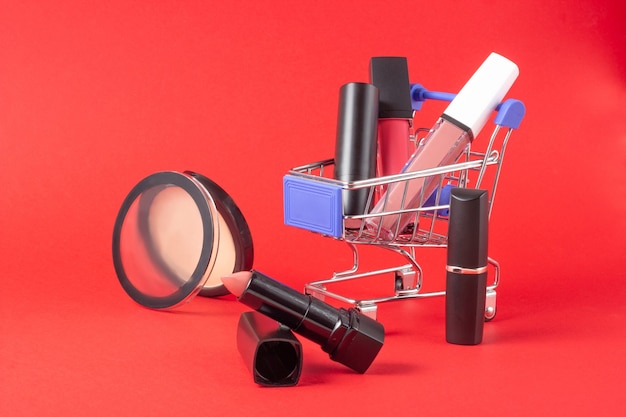 A set of lipstick cosmetics and lip glosses, powder, eyeshadow and a shopping trolley on a bright red background. The concept of buying cosmetics, online shopping, holiday