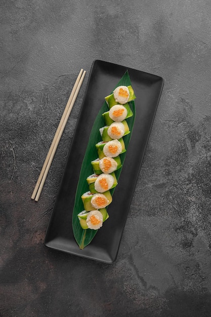 Set of maki rolls with cucumber salmon caviar and green bamboo leaf in a black ceramic plate with chopstick on a dark gray textured background top view