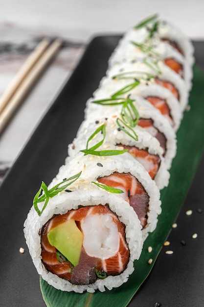 Set of maki rolls with salmon tuna shrimp avocado rice and green bamboo leaf in a black ceramic plate with chopstick on a bright white textured marble background side view closeup