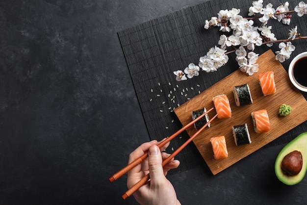 Фото Набор роллов суши и маки с нарезанным авокадо, рукой с палочками для еды и ветвью белых цветов на каменном столе. вид сверху.