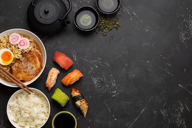 사진 일본 전통 음식과 음료 세트: 수프 라면, 삶은 쌀, 스시, 롤, 녹차는 검은 돌 배경의 찻주전자에 있습니다. 일본 요리, 위에서, 텍스트를 위한 공간