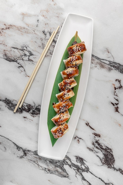 Set of philadelphia rolls with eel and green bamboo leaf in a white ceramic plate with chopstick on a bright textured marble background top view