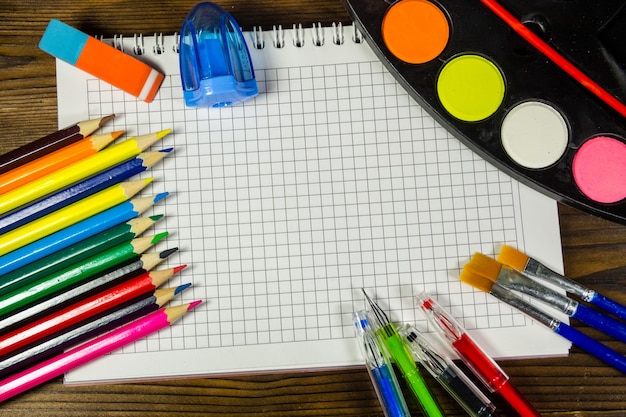 Set of school stationery supplies. Blank notebook, colored pencils, pens, watercolor paints and other items on wooden desk. Back to school concept