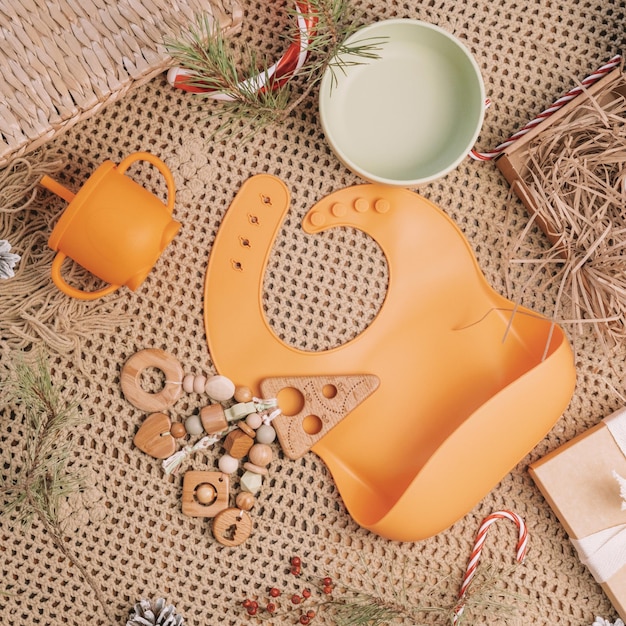 Set of silicone baby dishes, bib and wooden teether on beige blanket with Christmas decorations. Flat lay. Square frame