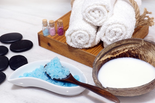 Set for Spa procedures from coconut oil, soft towels, hot stones and blue bath salt on a white marble table.