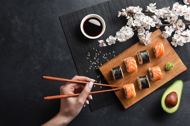 Set of sushi and maki rolls, sliced avocado, hand with chopsticks and branch of white flowers on stone table. Top view.