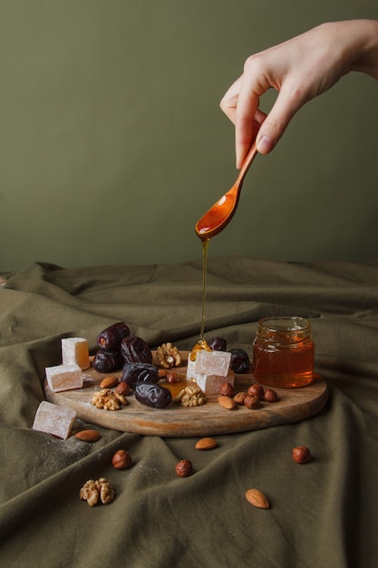 Set for Tea drinking. Hand holding spoon with dripping honey. Various sweets, nuts and honey for tea on a wooden cutting board. Healthy sweets, delicious dessert, natural sweets.