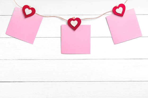 Set of three blank stickers hanging on string with clothespins behind white wooden surface.  Paper notes held on rope with red hearts. Copy space, top view.