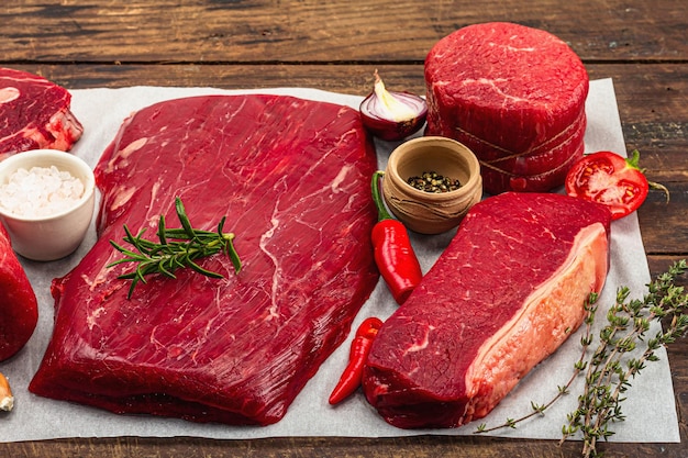 Set van verschillende steaks met traditionele specerijen en kruiden Vers rauw vlees omvat ribeye ronde flank en lendenbiefstuk Oude houten achtergrond plat lag close-up