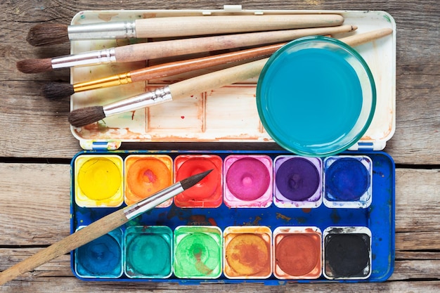 Set of watercolor paints art brushes and glass of water on old wooden table Top view