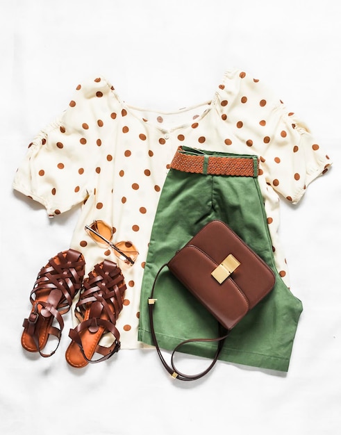 A set of women's summer clothes bermuda shorts polka dot blouse braided leather sandals and a cross body bag on a light background top view