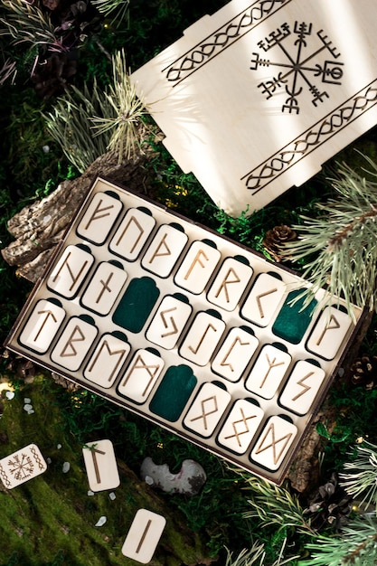 箱の中の木製のルーン文字のセットは、森の苔の上にあります。スカンジナビアのルーン文字が刻まれている長方形の木製のプラットフォームは、塩、円錐形、モミの針、樹皮に囲まれた緑の苔の上にあります