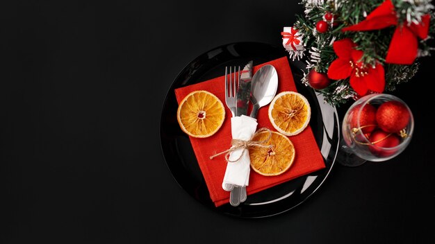 Фото Установка для праздничного рождественского ужина на черном столе с новогодним украшением и сухими апельсинами. красные елочные шары в бокале для вина