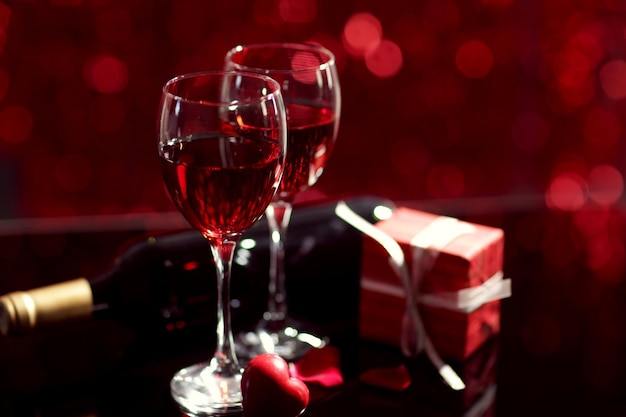 Impostazione di bicchieri con vino, bottiglia e un regalo nella scatola, su sfondo sfocato rosso