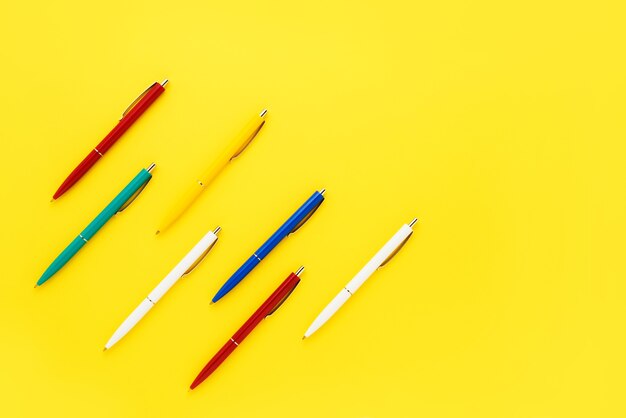 Seven colorful ballpoint pens on a bright yellow background.