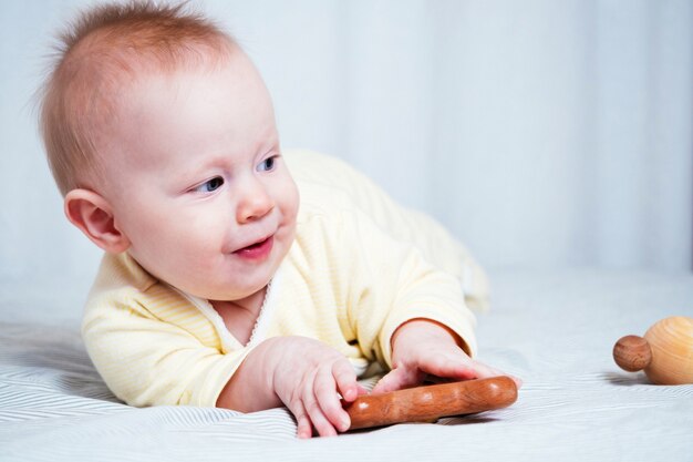 Una bambina di sette mesi è felice, sorride e gioca con i giocattoli di legno, sdraiata a pancia in giù in una stanza luminosa.