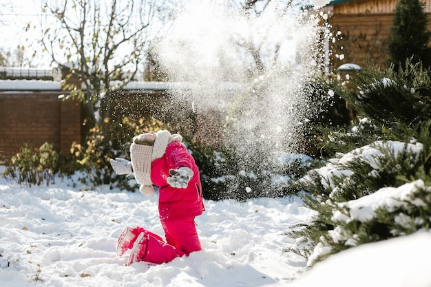 Seven-year cute girl in winter clothes playing with snow in the backyard of a house on a winter sunny day.