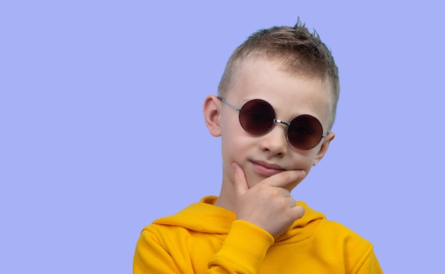 A seven-year-old boy in a yellow hoodie poses in front of a camera on a blue background