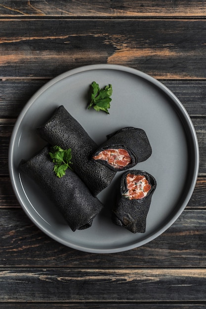 Several black cuttlefish ink pancakes with salmon and cheese filling on a gray plate