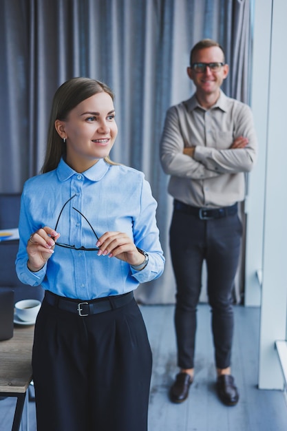 Several friendly colleagues are standing by the office window A business woman in glasses stands on the background of a man in a shirt Selective focus Portrait of business people