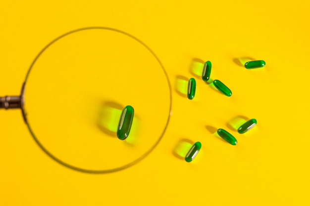 虫眼鏡の下の黄色の上にいくつかの緑色の透明なカプセルが配置されています。薬理学の。