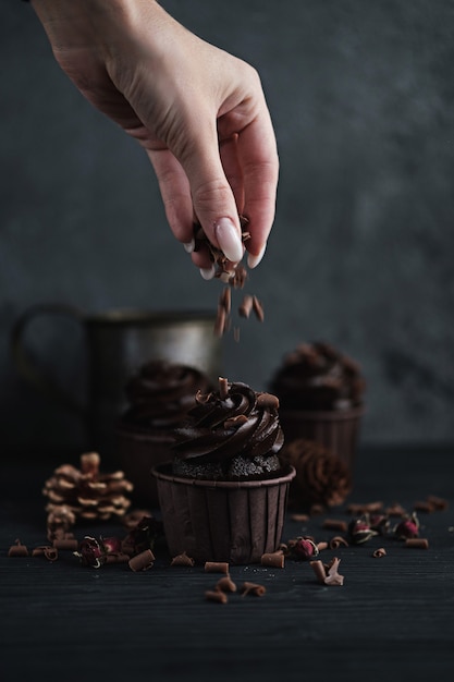 Several muffins or cupcakes with chocolate shaped cream at black table. Festive candle burns on a chocolate cake. A woman's hand crumbles grated chocolate onto a cake.