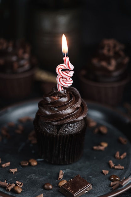 Several muffins or cupcakes with chocolate shaped cream at black table. Festive candle burns on a chocolate cake