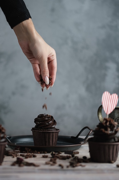 白いテーブルにチョコレートの形をしたクリームといくつかのマフィンまたはカップケーキ。聖バレンタインデーのハートの形をしたカード。女性の手がすりおろしたチョコレートをケーキに砕きます。