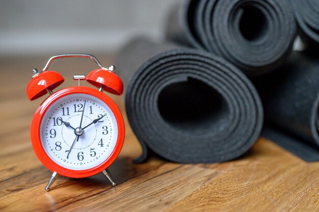 Several rolled yoga or fitness rubber mats black color and red alarm clock on wooden floor. sport accessories.