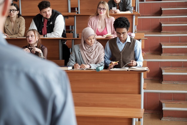 Several rows of contemporary intercultural university students\
sitting by long wooden desks in lecture hall and listening to\
professor at lesson