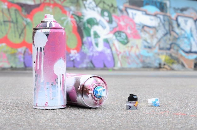 Several used spray cans with pink and white paint and caps for\
spraying paint under pressure is lies on the asphalt near the\
painted wall in colored graffiti drawings