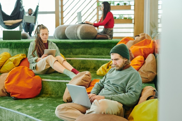 Several young intercultural managers of large contemporary open space office using laptops while sitting against cushions on green floor