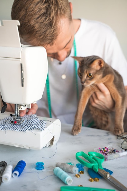 애완동물을 위한 옷 재봉 고양이를 위한 옷 재봉 미소 짓는 솔기가 아비시니안 고양이를 위한 옷을 바느질하고 있습니다. 작고 DIY 사업의 개념 진정한 감정 라이프스타일 선택적 초점