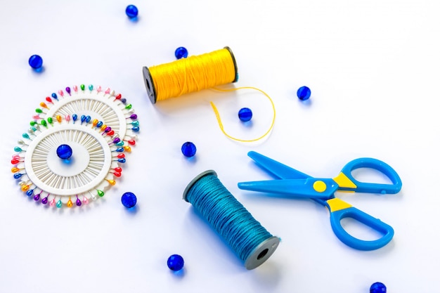 Sewing spools of blue and yellow threads, scissors, glass beds and colorful pins on white background