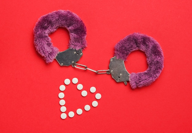 Sex stimulation. Handcuffs for sex games with pills (form heart) on red background. Sexual bdsm toy. Love, erotic concept. Top view