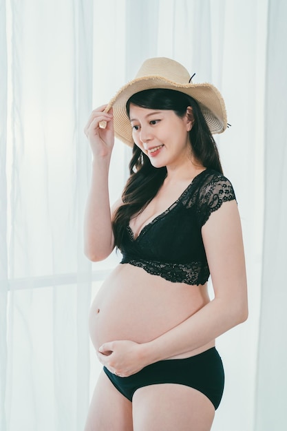 Sexy asian korean pregnant woman with long hair and straw hat on white background. Fashion studio portrait. young female parenthood in black lingerie and panties with bare belly going on vacation.