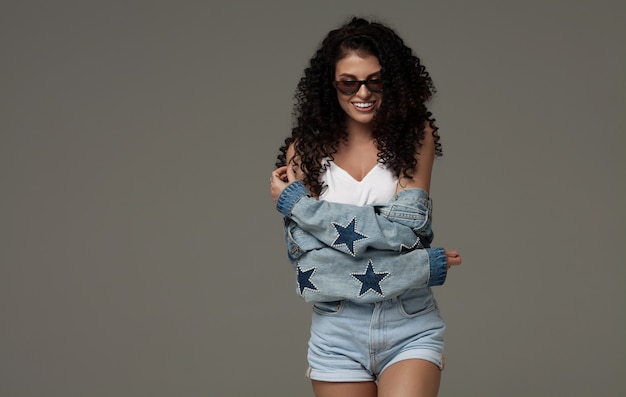 Sexy attractive brunette woman posing in fashionable jeans in studio. Fashion model posing. Long curly hair.