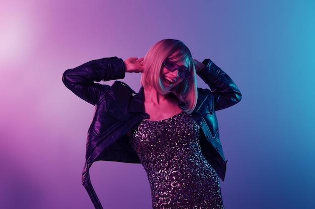Sexy beautiful blonde lady in leather jacket sparkly dress\
trendy sunglasses dance hold hands up posing isolated in blue pink\
color light studio background neon party cyberpunk concept copy\
space