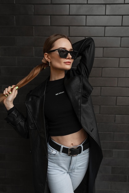 Sexy beautiful fashionable woman model in stylish black street clothes with a black leather long jacket Tshirt jeans and sunglasses standing near a black brick wall