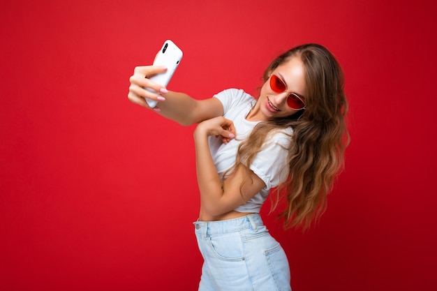 Sexy beautiful young blonde woman holding mobile phone taking selfie photo using smartphone camera