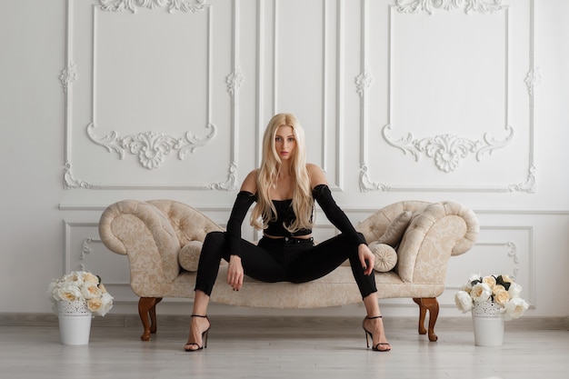 Sexy beautiful young model woman in fashionable black clothes with jeans sitting on a sofa in a vintage room