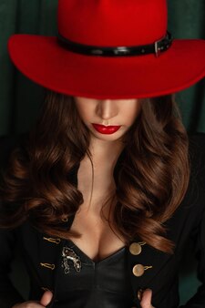 Bella giovane donna sexy con acconciatura di labbra, cappello e riccioli rossi in blazer nero alla moda con seni su uno sfondo verde vintage. ritratto sensuale femminile di fascino