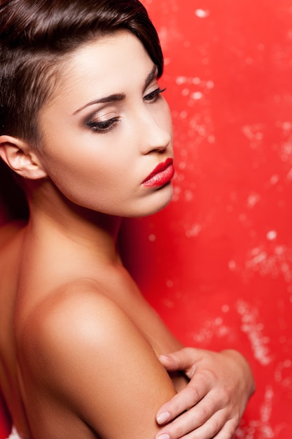 Sexy beauty. Side view of beautiful young shirtless woman standing against red background and covering breasts with hands