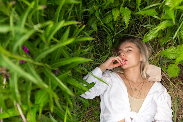 Sexy blonde girl in a white shirt with red lipstick and red manicure lies in the green grass.