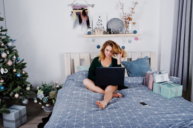 Sexy blonde model in green lingerie on bed with black laptop against new year tree.