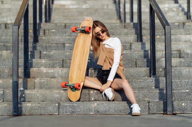 Сексуальная блондинка с лонгбордом сидит на лестнице в коротких шортиках. Фото высокого качества