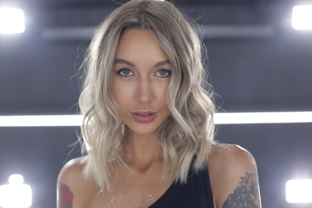 Sexy blonde with sequins around her neck stands indoors with bright light and sensually looks at the camera high quality photo