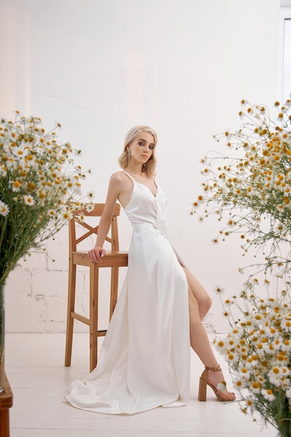 A sexy blonde woman in a beautiful white dress is standing at home among bouquets of wild flowers