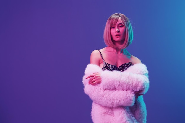 Sexy blonde woman in fluffy fur coat sparkly dress with open shoulders cross hands posing isolated in blue violet pink color light studio background Neon party Fashion concept Copy space Banner