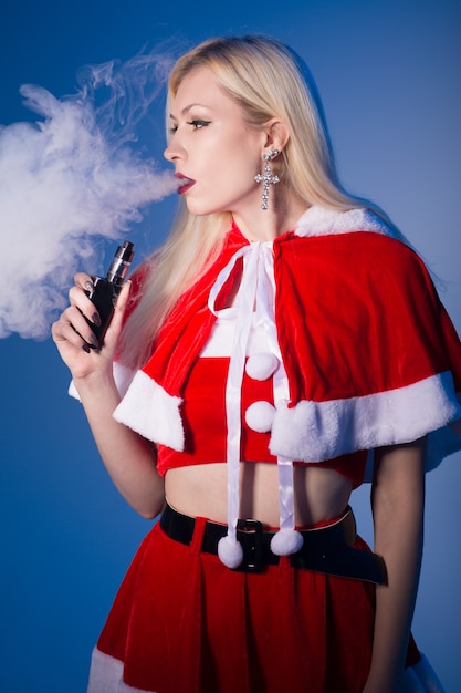 Foto donna bionda sexy in costume da babbo natale che fuma sigaretta elettronica ed espira vapore